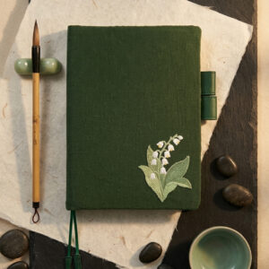 A forest green A5 linen book cover with embroidered white lily of the valley flowers on handmade paper and black slate with a calligraphy brush and tea cup