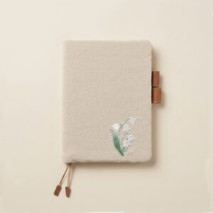 Minimalist flatlay of a handcrafted A5 natural oatmeal linen journal cover with delicate embroidered white lily of the valley flowers, tan leather pen loops, and twin bookmark cords on a neutral background.