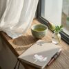 Handcrafted taupe linen notebook cover with lotus embroidery on a rustic wooden windowsill with a tea bowl, a bud in a vase, and a pen under natural sunlight.