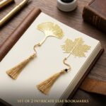 A set of two gold metal hollow gingko and maple leaf bookmarks with tassels resting on an open journal on a dark wooden desk.