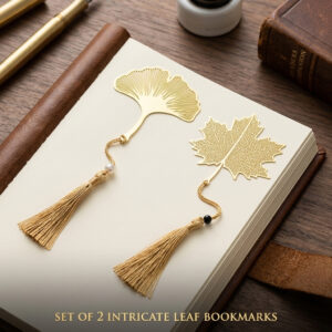 A set of two gold metal hollow gingko and maple leaf bookmarks with tassels resting on an open journal on a dark wooden desk.